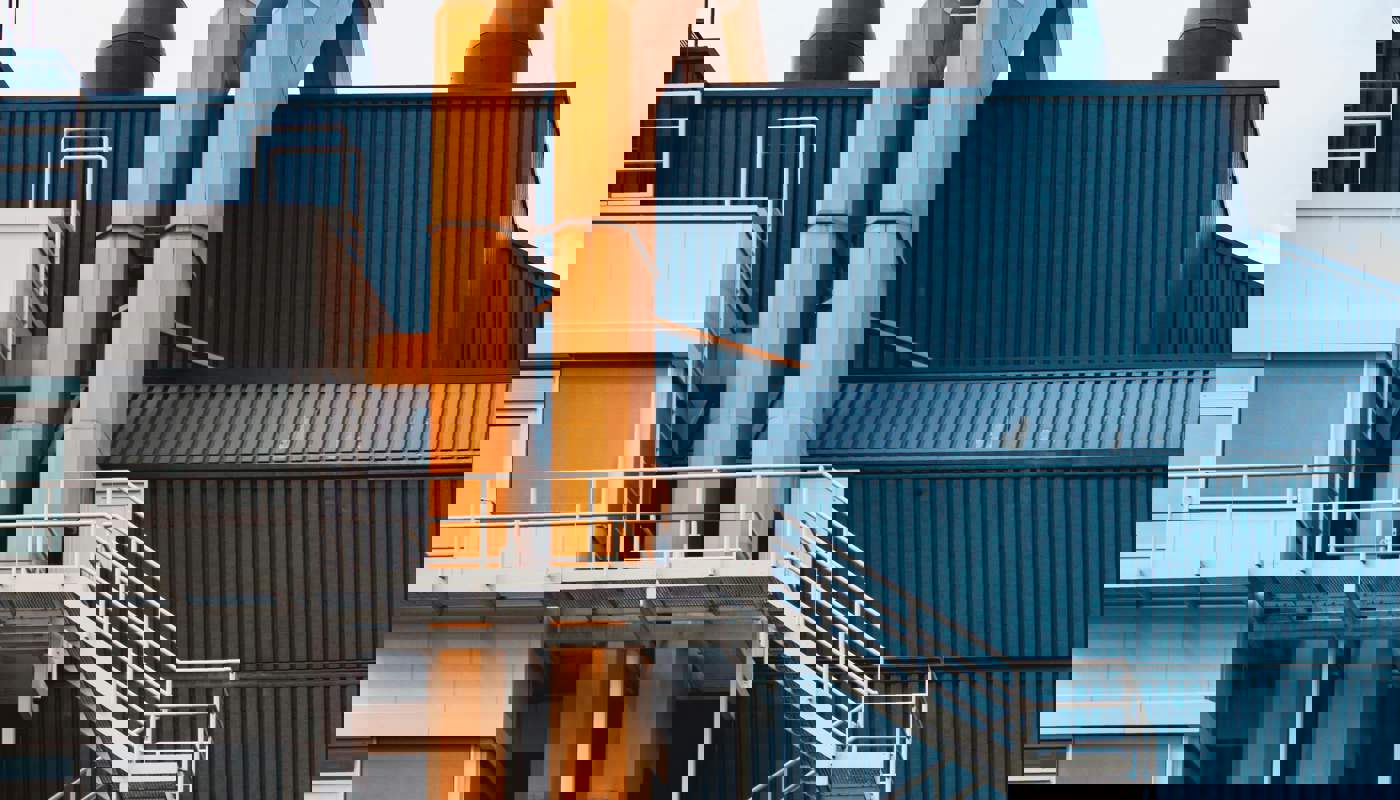 Close-up view of a contemporary industrial building featuring blue corrugated metal panels, bright orange structural pipes, and white metal staircases on the exterior, set against a partly cloudy sky.
