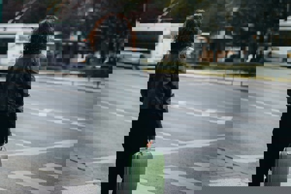 A person stands at a zebra crossing holding a green handbag, facing an empty road with a bus in the distance and trees in the background. The image symbolises being at a crossroads, reflecting the consequences of concealing secondary employment and the uncertainty that follows dismissal.