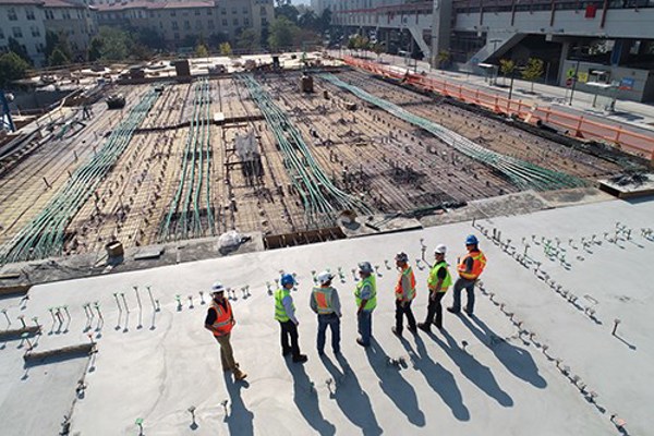 Construction workers in safety vests and helmets standing on a concrete surface at an early-stage construction site, with green pipes, and orange safety barriers visible in the background.