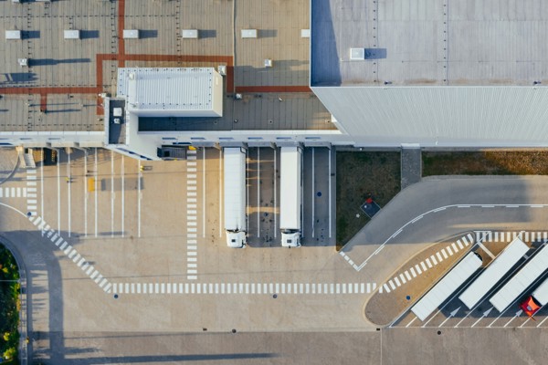 Aerial view of a modern industrial facility with loading docks and parked lorries. The image shows two white trucks positioned at loading bays, a large warehouse building with a grey roof, and marked lanes for vehicle movement.