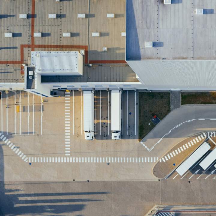 Aerial view of a modern industrial facility with loading docks and parked lorries. The image shows two white trucks positioned at loading bays, a large warehouse building with a grey roof, and marked lanes for vehicle movement.