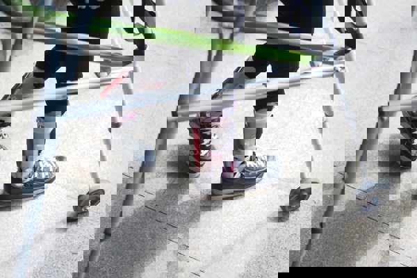 Close-up of a person using a metal walker for mobility, wearing black shoes and orthotic leg braces on both legs, walking on a tiled surface.