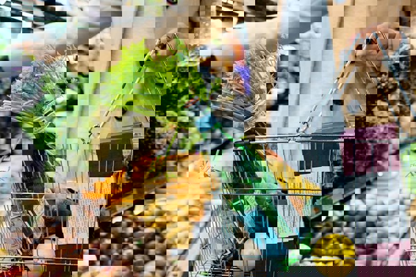 Person shopping in a grocery store, holding a metal basket filled with fresh produce including lemons, leafy greens, and bottled water, while selecting vegetables from a display of root crops and greens.