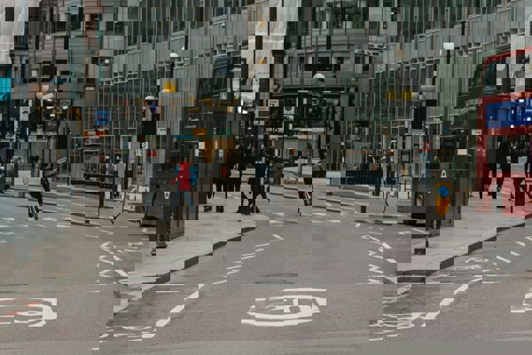 Cyclist riding through a city street junction with pedestrians crossing and a red double-decker bus beside modern office buildings.