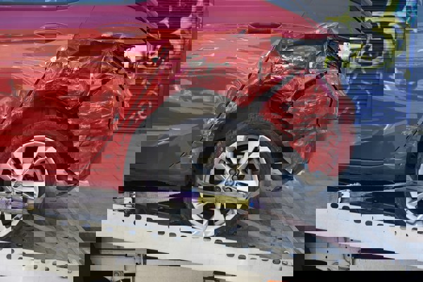 Side view of a red car with severe damage to the rear section, secured with straps on a recovery truck.